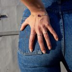 Złota biżuteria autorska, pierścionek z diamentami oraz bransoletka z kolorowymi kamieniami na modelce, która trzyma rękę na niebieskich spodniach.